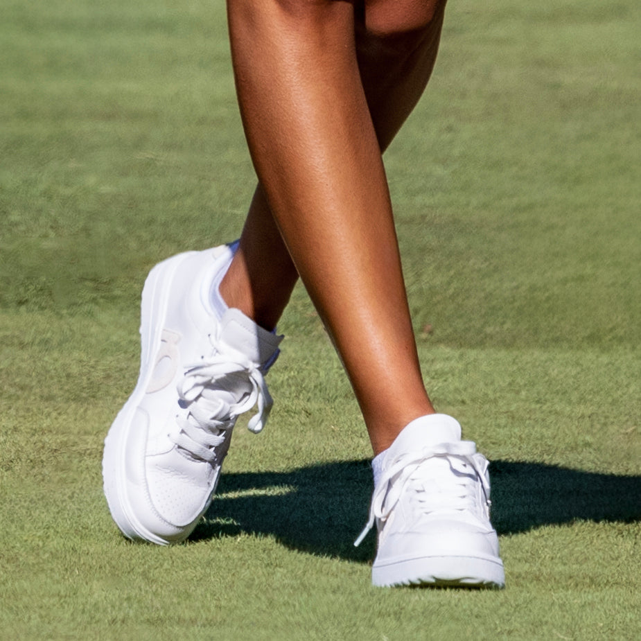 White low-top athletic sneakers worn on a person's feet, shown in a close-up outdoor shot on a green grass surface.