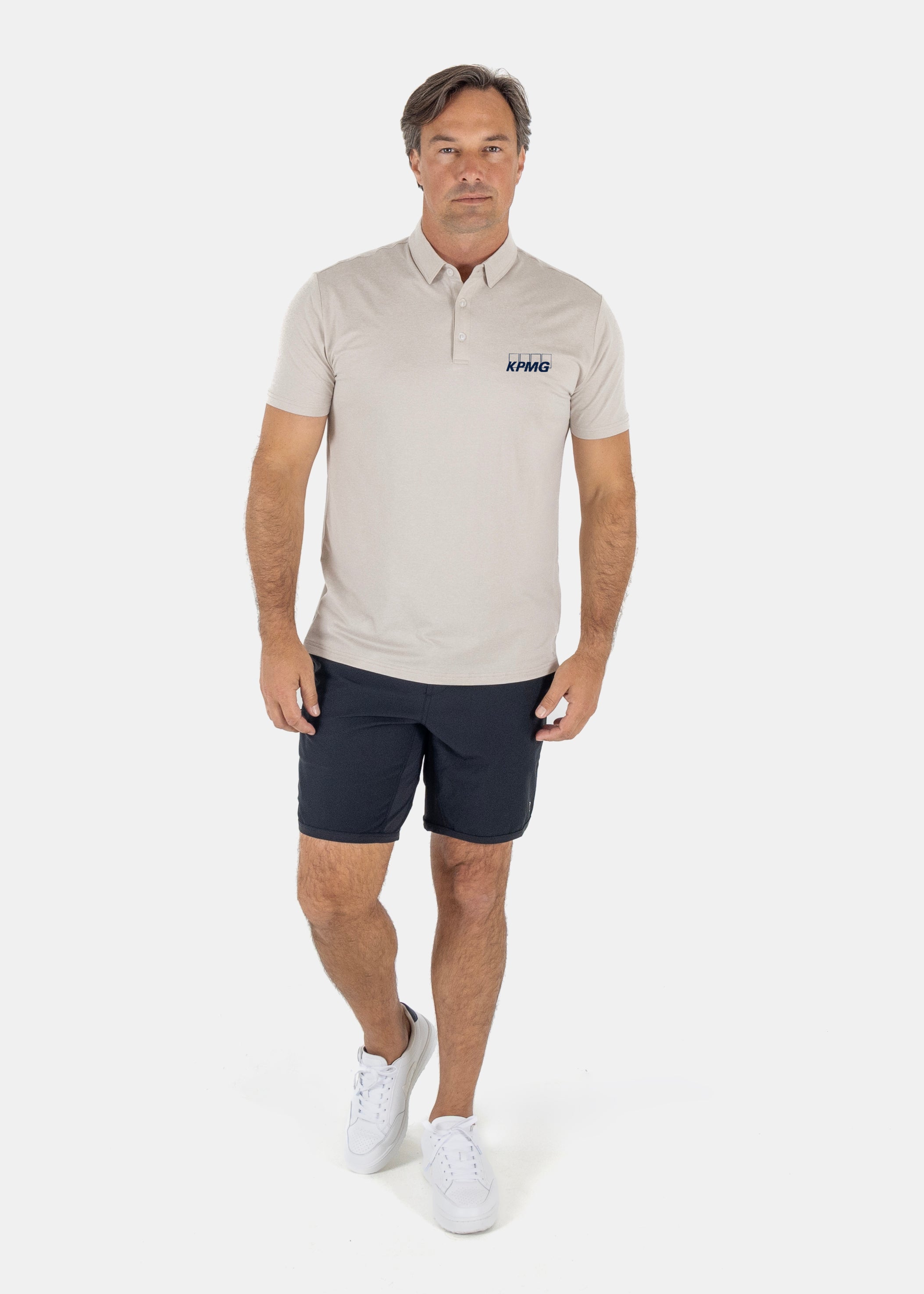 A man modeling a light beige short-sleeve polo shirt with a small navy logo on the left chest, paired with khaki shorts and white sneakers, against a plain white studio background.