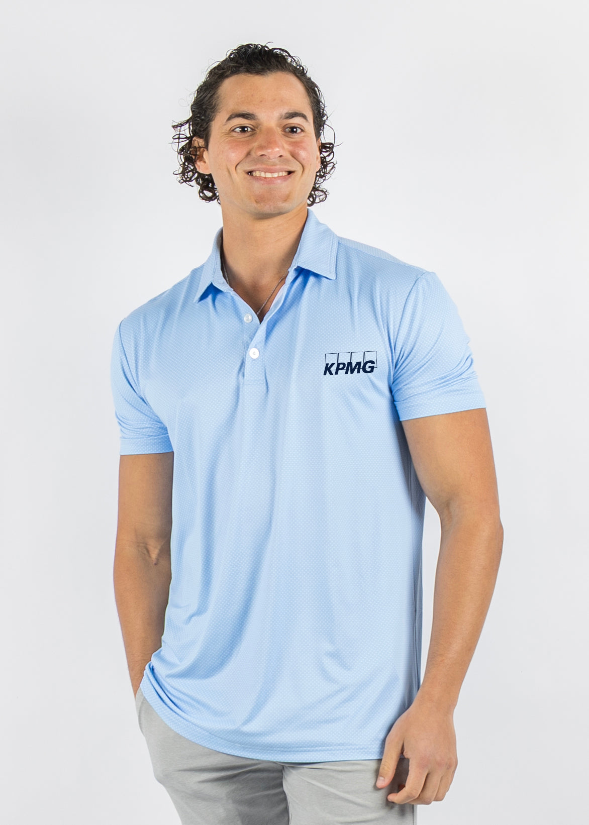 A man wearing a light blue short-sleeve polo shirt with a black KPMG logo on the left chest, paired with light gray trousers, photographed against a plain light gray studio background.
