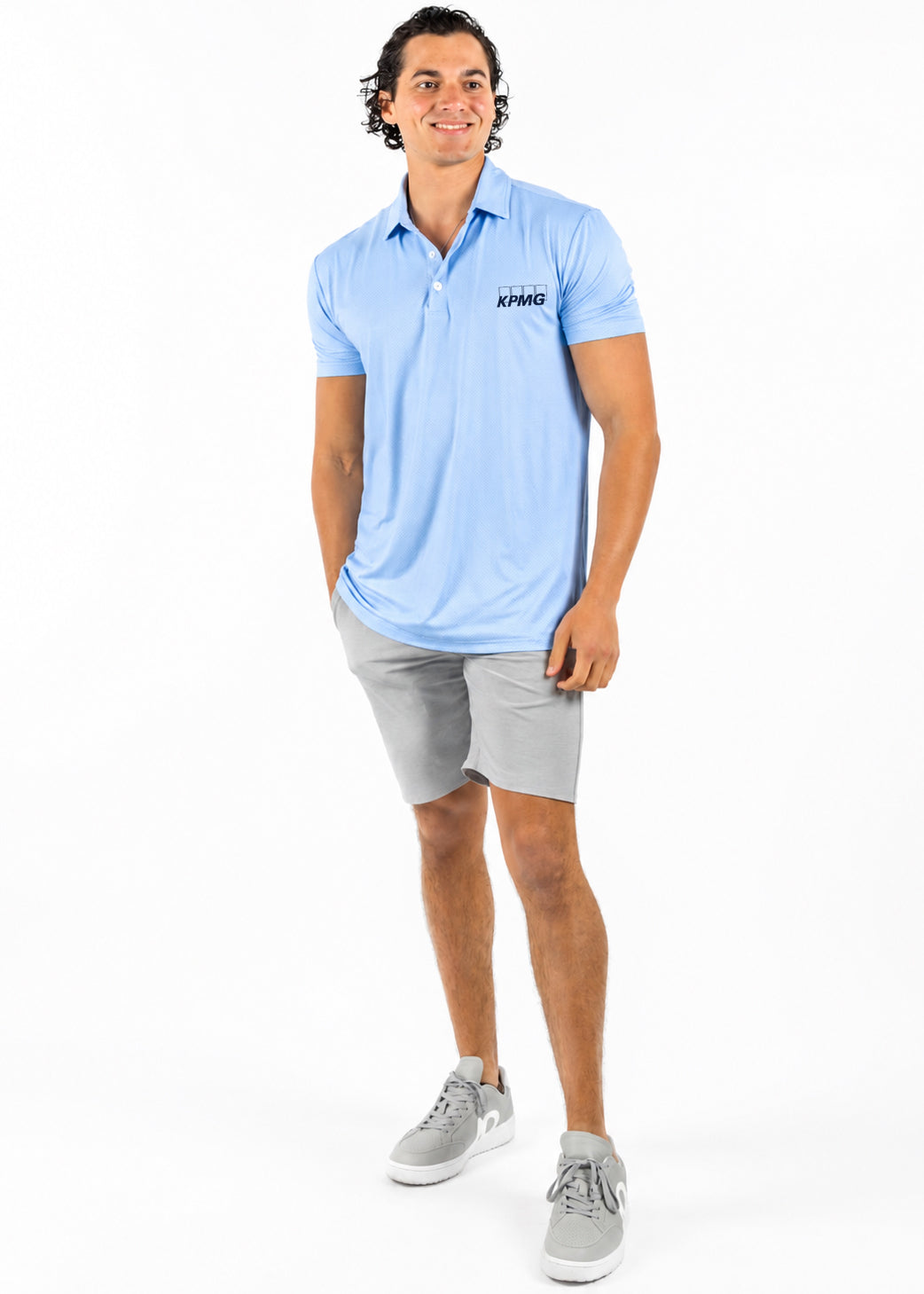 A man modeling a light blue short-sleeve polo shirt with a small black 'KYMEX' logo on the left chest, paired with gray shorts and gray sneakers, against a plain white background.