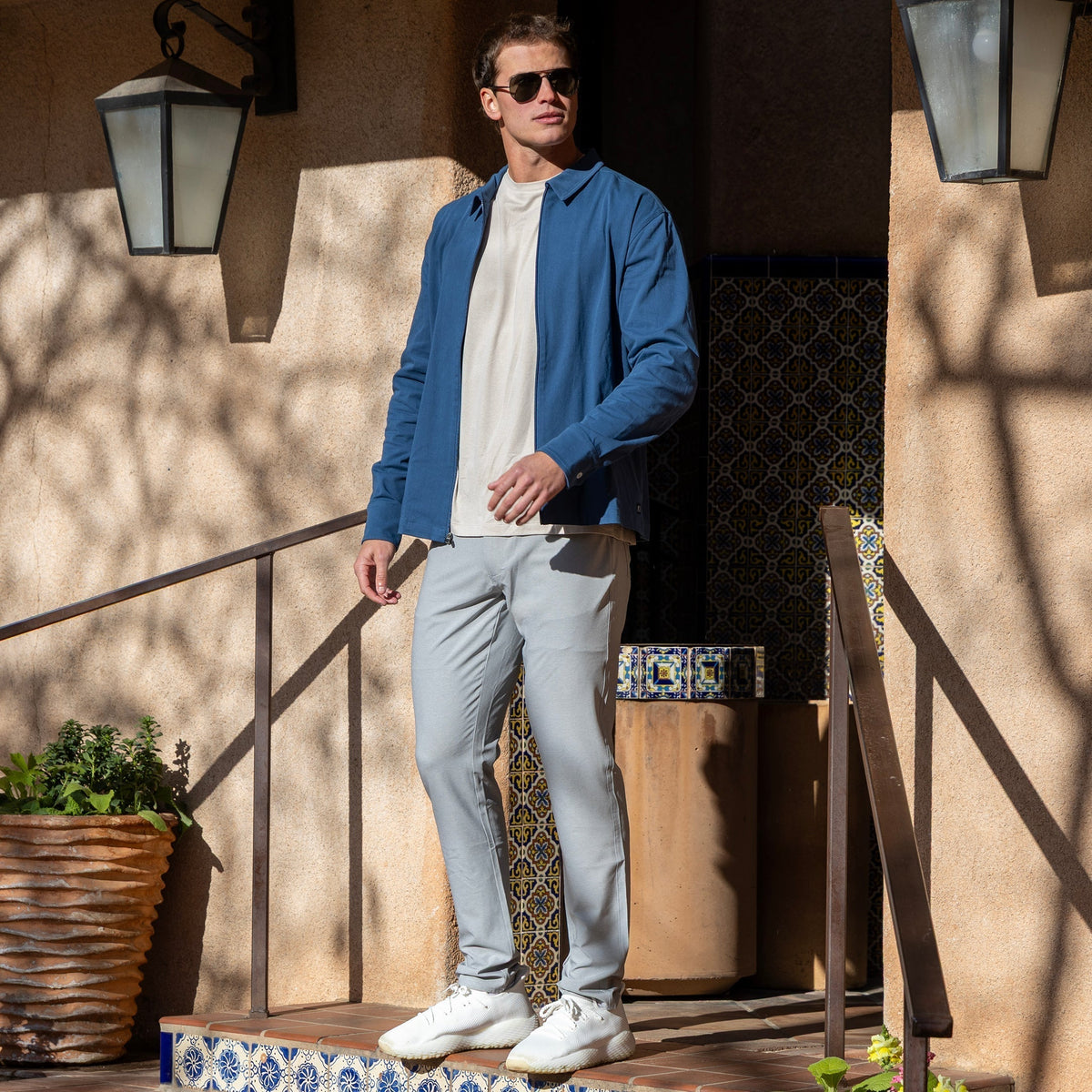 Man wearing a blue jacket over a light T-shirt with gray pants, white sneakers, and sunglasses on sunlit tiled steps outside a stucco building.