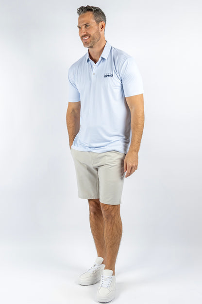 A man modeling a light blue short-sleeve polo shirt with a small dark logo on the left chest, paired with beige shorts and white sneakers, against a plain white studio background.