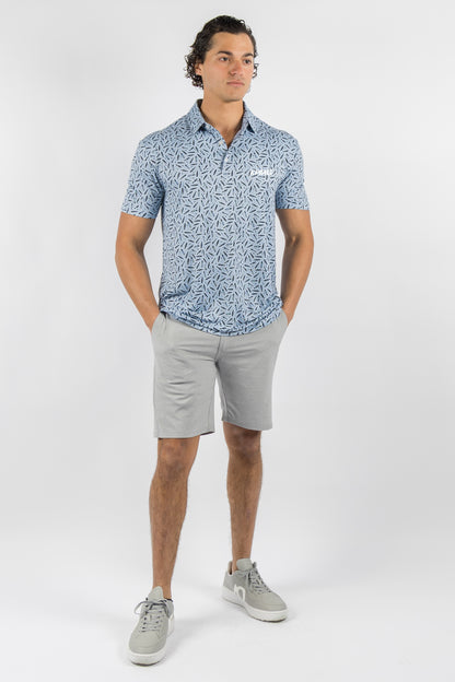 A man modeling a short-sleeve polo shirt with a blue and white abstract pattern, paired with light gray shorts and gray sneakers, standing against a light gray studio background.