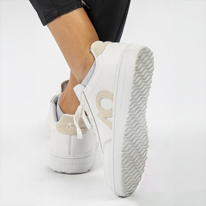 White low-top sneakers with beige accents, shown on a model's feet against a clean white studio background.