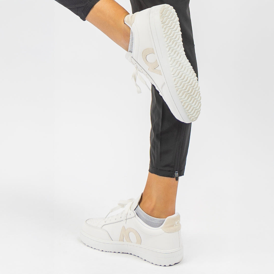 White low-top sneakers with beige AO logo, shown on a model wearing black pants against a clean white studio background.