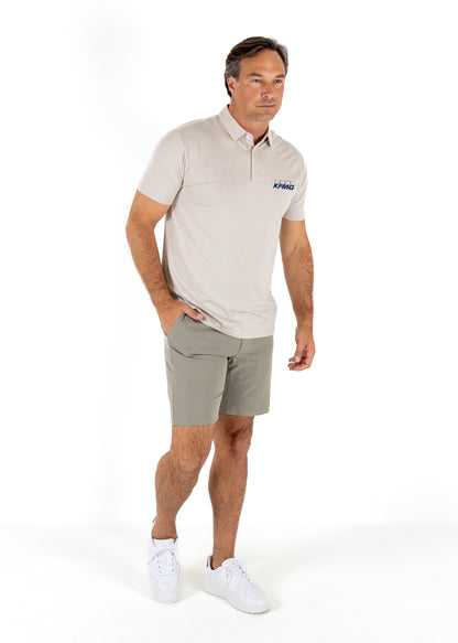 A man modeling a light beige short-sleeve polo shirt with a small navy logo on the left chest, paired with khaki shorts and white sneakers, against a plain white studio background.