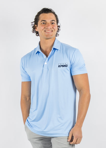 A man wearing a light blue short-sleeve polo shirt with a black KPMG logo on the left chest, paired with light gray trousers, photographed against a plain light gray studio background.