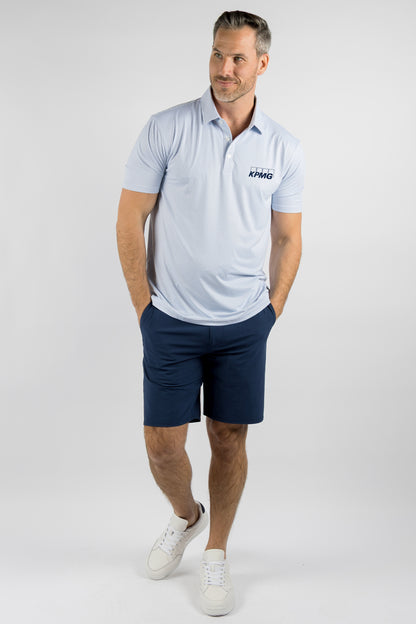A man modeling a light blue short-sleeve polo shirt with a dark blue logo on the left chest, paired with navy blue shorts and white sneakers, against a light gray studio background.