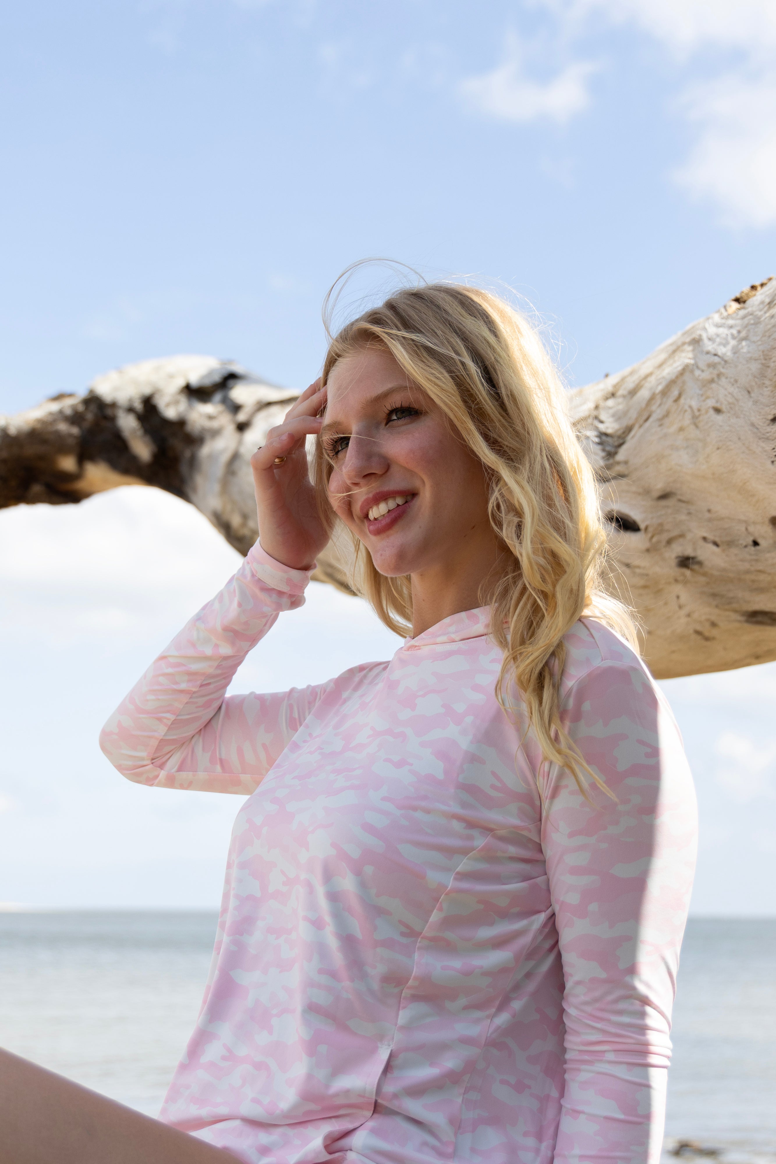 Woman in a pink camo hoodie standing on a beach with driftwood in the background